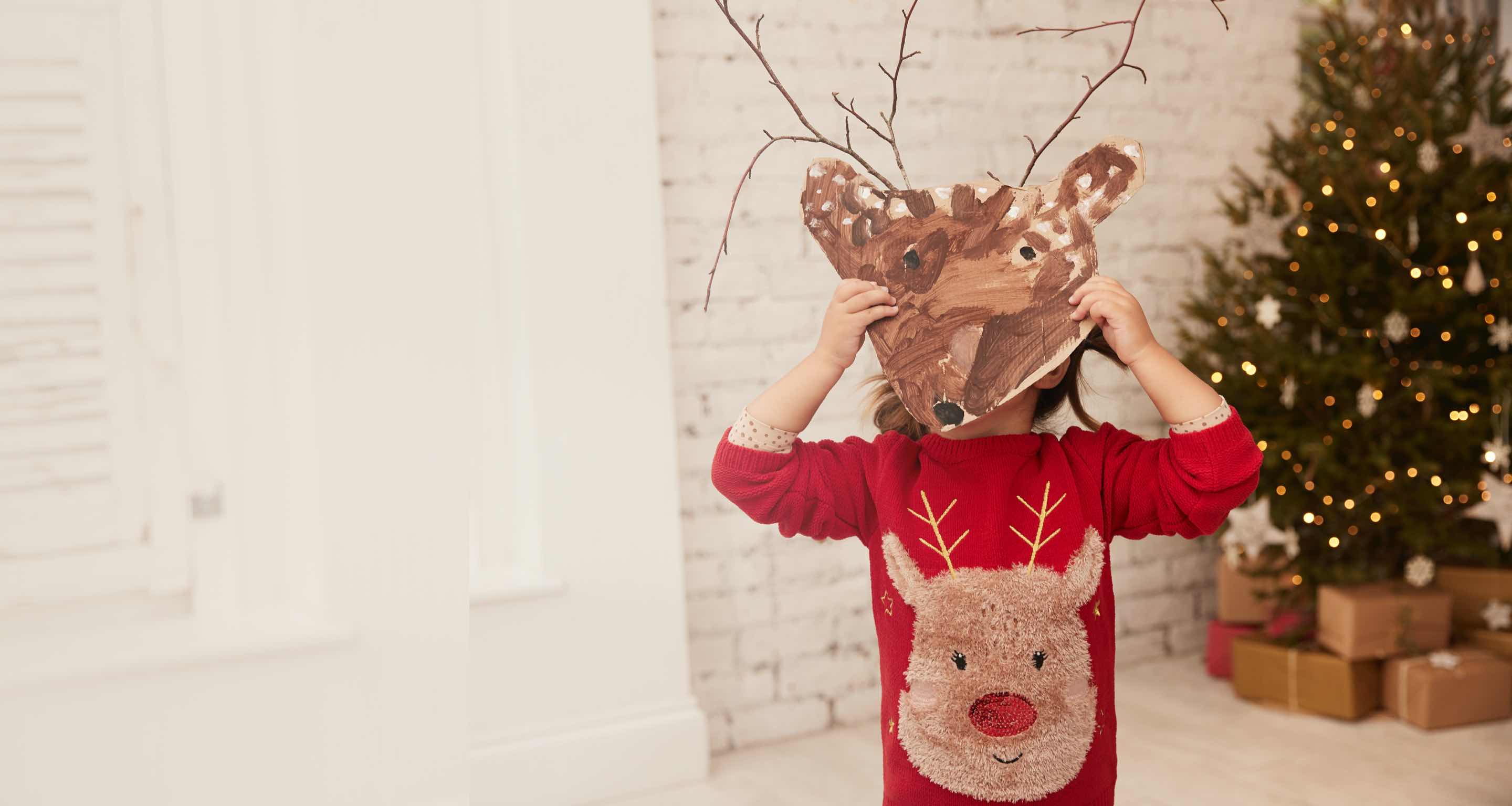 Little girl wearing a red reindeer fluffy jumper holding up a hand painted reindeer mark with twigs for antlers.